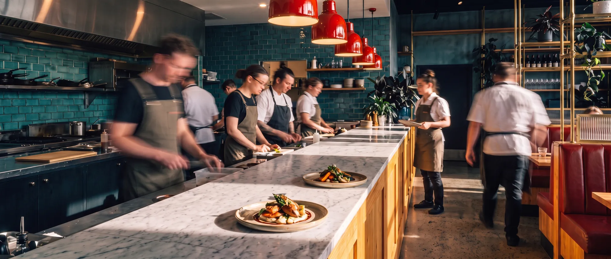 Kuchaři a obsluha v pohybu za barem restaurace, v popředí talíř a sklenka vína
